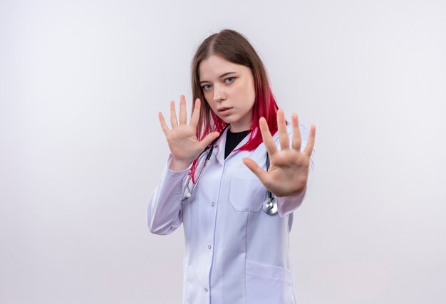 młoda kobieta lekarz noszenie szaty medycznej stetoskop pokazując gest zatrzymania na odizolowanej białej ścianie
