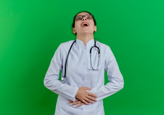 Młoda kobieta lekarz ubrana medyczną szatę i stetoskop i okulary patrząc w górę trzymając brzuch i śmiejąc się na białym tle na zielonej ścianie z miejsca na kopię