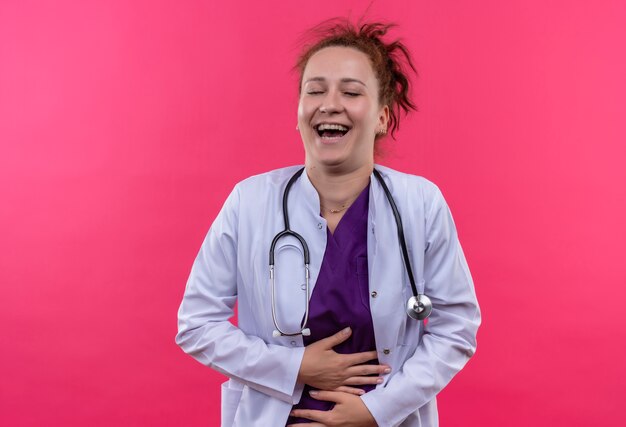 Młoda Kobieta Lekarz Ubrana W Biały Fartuch Ze Stetoskopem, śmiejąc Się, Dotykając Jej Brzucha Stojącego Na Różowej ścianie
