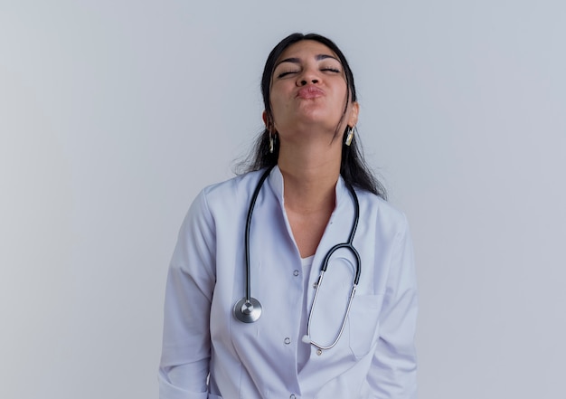 Młoda kobieta lekarz ubrana w medyczny szlafrok i stetoskop robi gest pocałunku z zamkniętymi oczami na białym tle