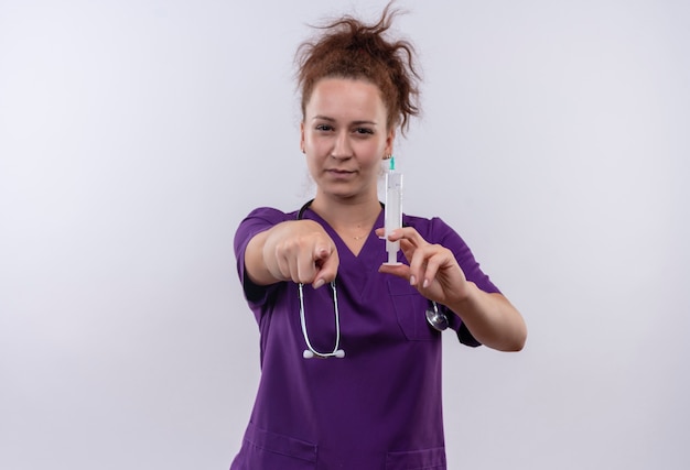 Bezpłatne zdjęcie młoda kobieta lekarz ubrany w mundur medyczny ze stetoskopem trzymając strzykawkę wskazując palcem patrząc pewnie stojąc na białej ścianie