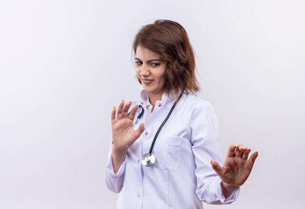 Młoda kobieta lekarz w białym fartuchu ze stetoskopem czyniąc gest obrony rękami z obrzydzonym wyrazem stojącym na białej ścianie