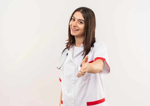 Młoda kobieta lekarz w białym fartuchu ze stetoskopem na szyi patrząc do przodu uśmiechnięta przyjazna oferująca dłoń stojąca nad białą ścianą