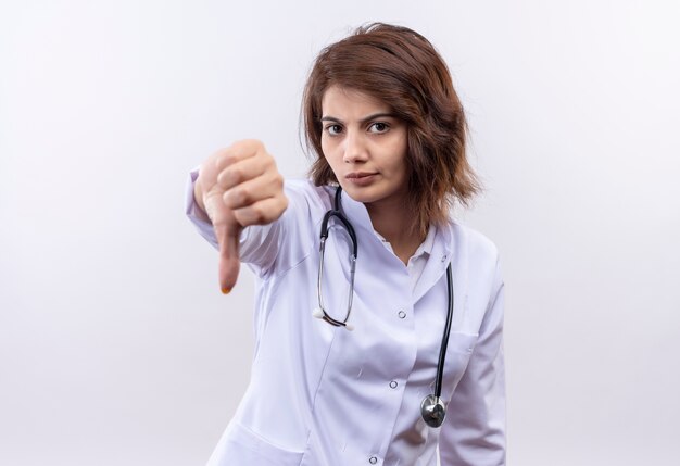Młoda kobieta lekarz w białym fartuchu ze stetoskopem patrząc na kamery niezadowolony pokazując kciuk w dół