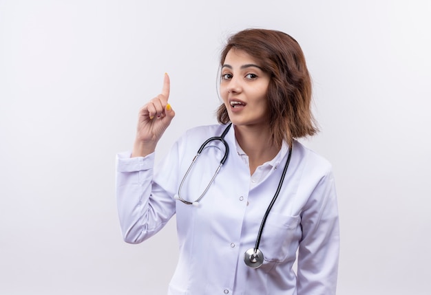 Młoda kobieta lekarz w białym fartuchu ze stetoskopem patrząc na kamery uśmiechnięta pewnie wskazując z finfer