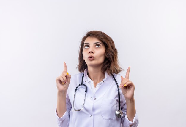 Młoda kobieta lekarz w białym fartuchu ze stetoskopem patrząc zaskoczony, wskazując na finfers stojąc nad białą ścianą