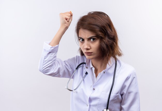 Młoda kobieta lekarz w białym fartuchu ze stetoskopem podnosząc pięść z gniewną twarzą stojącą na białej ścianie