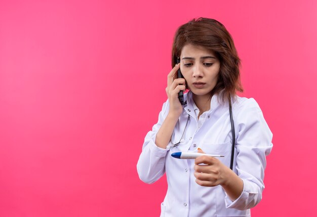 Młoda kobieta lekarz w białym fartuchu ze stetoskopem trzymając cyfrowy termometr patrząc na to martwi się podczas rozmowy przez telefon komórkowy