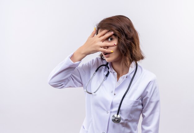 Młoda kobieta lekarz w białym fartuchu ze stetoskopem zaskoczona i zmartwiona zasłaniająca oczy dłońmi patrząc przez palce stojące na białej ścianie