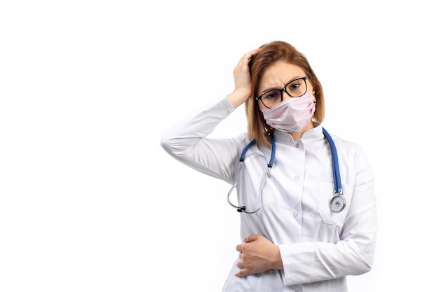 młoda kobieta lekarz w białym garniturze medycznym ze stetoskopem w białej masce ochronnej pozowanie na biały