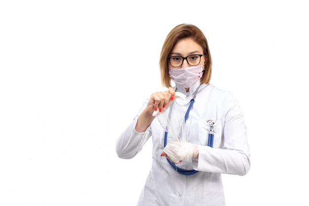Bezpłatne zdjęcie młoda kobieta lekarz w białym garniturze medycznym ze stetoskopem w białej masce ochronnej ustalające biały bandaż na białym