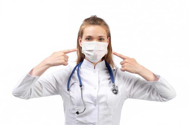 młoda kobieta lekarz w białym kombinezonie medycznym w stetoskop białej maski ochronnej wskazując palcami na biały