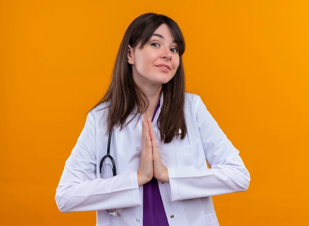 Młoda kobieta lekarz w medycznej szacie ze stetoskopem składa ręce na odizolowanych pomarańczowej ścianie
