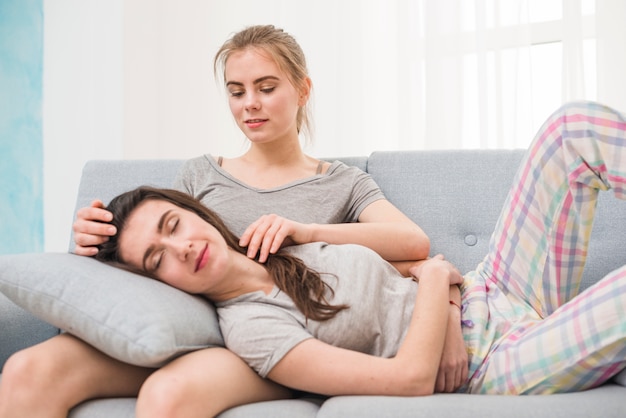 Bezpłatne zdjęcie młoda kobieta lesbijek spanie na kolanach jej dziewczyny siedzi na kanapie w domu