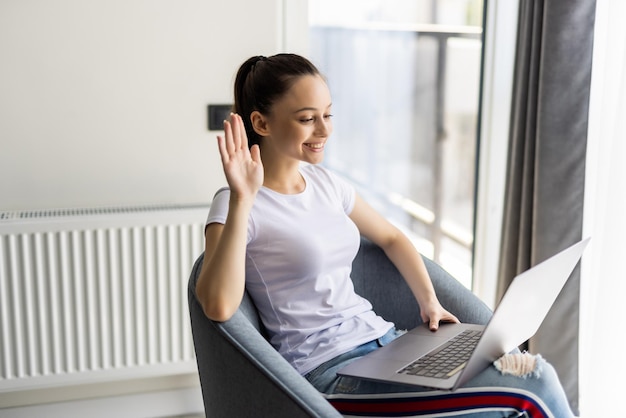 Bezpłatne zdjęcie młoda kobieta macha ręką wideorozmowa laptop siedzieć wiklinowym krześle nosić ubrania w stylu casual w domu w pomieszczeniu