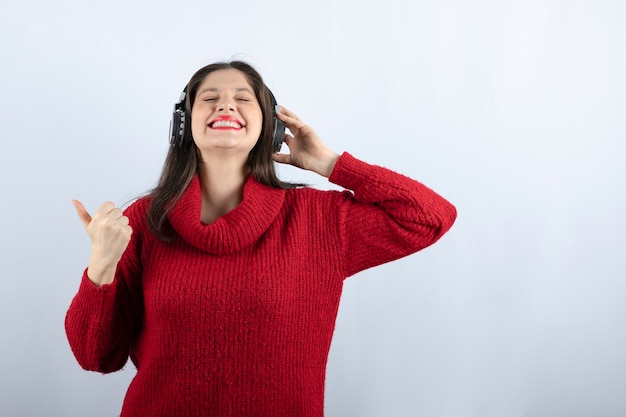 Młoda kobieta model w czerwonym swetrze ze słuchawkami pokazującymi kciuk w górę