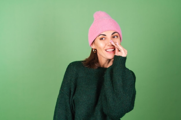 Młoda kobieta na zielono w ciepłym przytulnym swetrze i różowej czapce opowiada sekret, szepcze, plotkuje