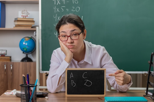 Młoda kobieta nauczyciel w okularach trzyma małą tablicę wskazującą palcem wskazującym, wyjaśniając lekcję, patrząc niezadowolony, siedząc przy szkolnej ławce przed tablicą w klasie