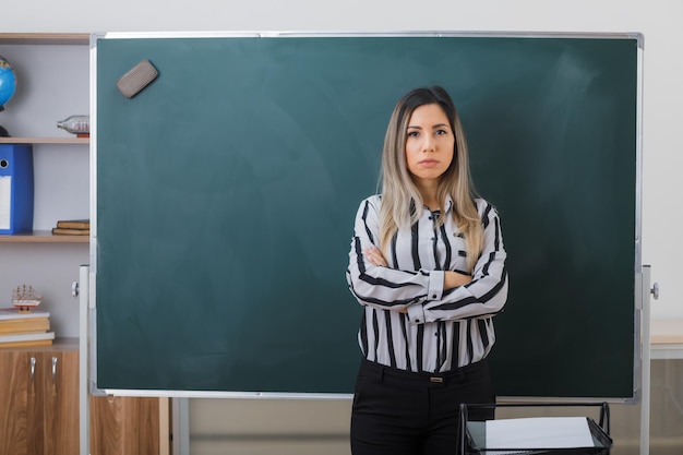 Bezpłatne zdjęcie młoda kobieta nauczycielka stojąca przy tablicy w klasie wyjaśniająca lekcję krzyżującą ręce na piersi, patrzącą na kamerę z poważną miną