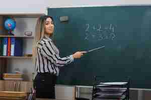 Bezpłatne zdjęcie młoda kobieta nauczycielka stojąca przy tablicy w klasie wyjaśniająca lekcję trzymającą wskaźnik uśmiechający się pewnie