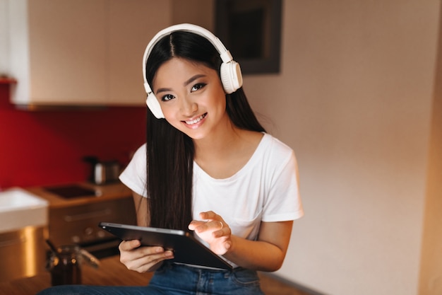 Młoda kobieta o ciemnych włosach z uśmiechem patrzy do przodu, trzyma tablet i słucha muzyki w słuchawkach