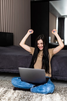 Młoda kobieta odrabiała pracę domową na podłodze, używając laptopa z szczęśliwym gestem wygranej i trzymając ręce w górze