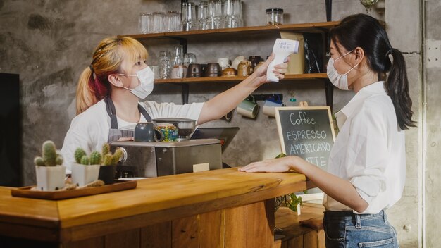 Młoda kobieta personelu restauracji nosząca maskę ochronną za pomocą termometru na podczerwień lub pistoletu temperaturowego na czole klienta przed wejściem.