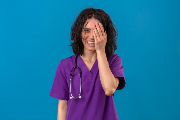 Młoda kobieta pielęgniarka w mundurze medycznym i ze stetoskopem, uśmiechając się i śmiejąc się z ręką na twarzy obejmującej oko na stojącą niespodziankę
