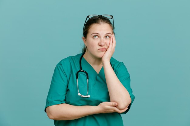 Młoda kobieta pielęgniarka w mundurze medycznym ze stetoskopem na szyi wygląda na zmęczoną i znudzoną, przewracając oczami w górę, stojąc na niebieskim tle
