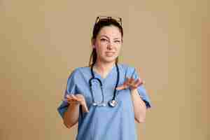 Bezpłatne zdjęcie młoda kobieta pielęgniarka w mundurze medycznym ze stetoskopem wokół szyi, patrząc na kamery, martwiąc się, co gest stop, rozciąganie rąk, stojąc na brązowym tle