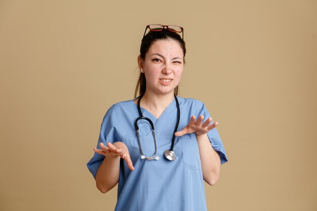 Młoda kobieta pielęgniarka w mundurze medycznym ze stetoskopem wokół szyi, patrząc na kamery, martwiąc się, co gest stop, rozciąganie rąk, stojąc na brązowym tle