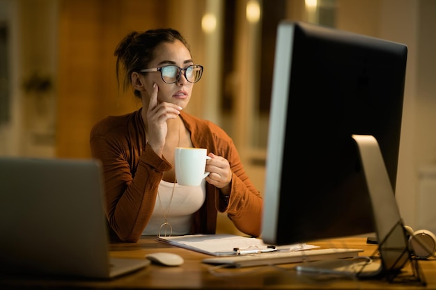 Młoda kobieta pijąca kawę i myśląca podczas pracy na komputerze stacjonarnym wieczorem w domu