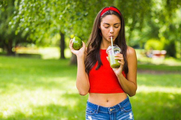 Młoda kobieta pije avocado smoothie w plastikowej filiżance przy outdoors