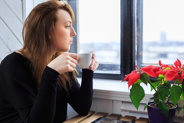 Młoda kobieta pije kawę i patrząc przez okno.