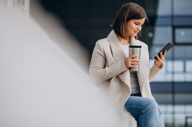 Młoda kobieta pije kawę i używa telefon na zewnątrz ulicy