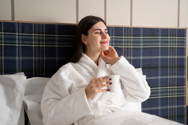 Młoda kobieta pije kawę siedząc na łóżku w pokoju hotelowym