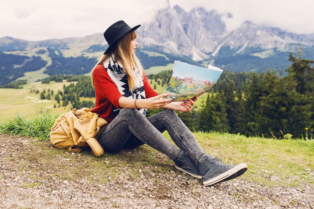 Młoda kobieta podróżnik z plecakiem i kapeluszem, siedząc na trawie i szukając właściwego kierunku na mapie