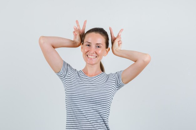 Młoda kobieta pokazująca znak v jako rogi w pasiastej koszulce i wyglądająca śmiesznie. przedni widok.