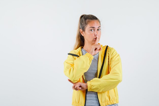 Młoda kobieta pokazuje gest ciszy w koszulce, kurtce i patrząc ostrożnie. przedni widok.