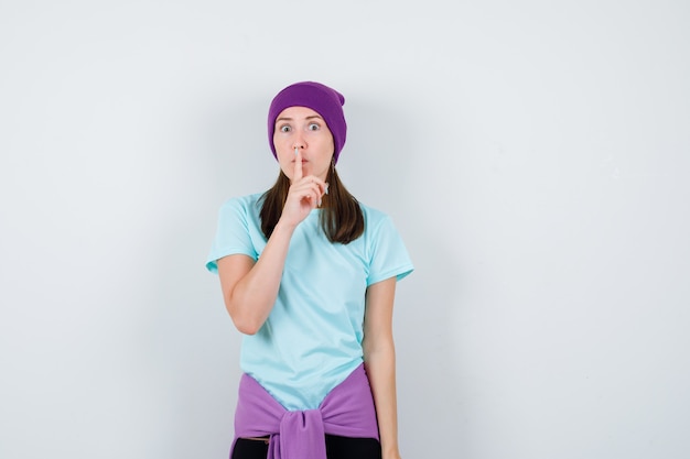 Młoda kobieta pokazuje gest ciszy w niebieskiej koszulce, fioletowej czapce i wygląda na przestraszoną. przedni widok.