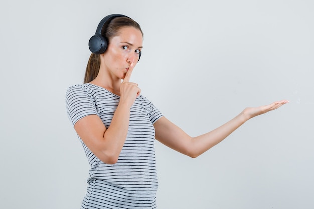 Młoda kobieta pokazuje gest ciszy z rozłożoną dłonią w t-shirt, słuchawki, widok z przodu.