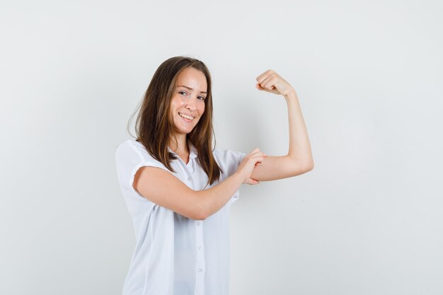 Młoda kobieta pokazuje mięśnie ramion w białej bluzce i wygląda potężnie