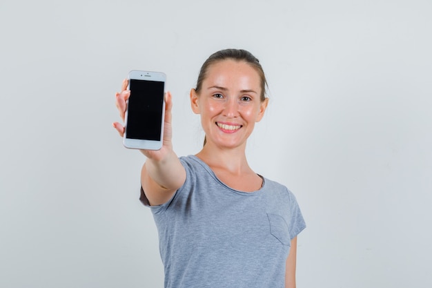 Młoda kobieta pokazuje telefon komórkowy w szarej koszulce i wygląda radośnie. przedni widok.