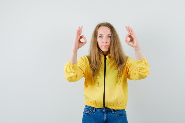 Młoda kobieta pokazuje znak ok obiema rękami w żółtej kurtce bomber i dżinsach i wygląda poważnie. przedni widok.