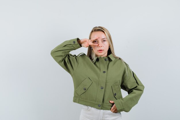Bezpłatne zdjęcie młoda kobieta pokazuje znak v nad okiem w zielonej kurtce, dżinsach i patrząc skoncentrowany. przedni widok.