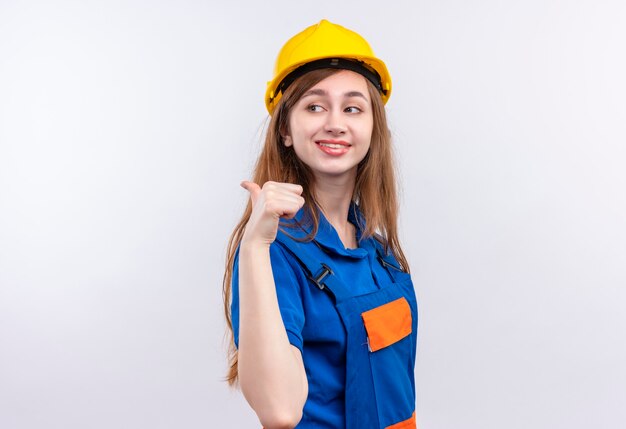 Młoda kobieta pracownik budowniczy w mundurze budowlanym i hełmie wskazującym kciukiem na bok uśmiechnięty pewny siebie stojący nad białą ścianą