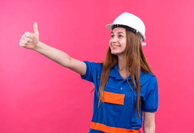 Młoda kobieta pracownik budowniczy w mundurze budowy i hełmie ochronnym patrząc na bok uśmiechnięty pokazując kciuki stojąc nad różową ścianą