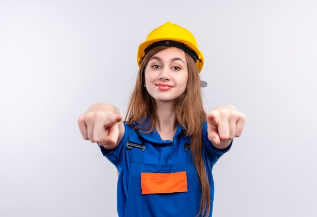 Młoda kobieta pracownik budowniczy w mundurze budowy i hełmie ochronnym patrząc pewnie uśmiechnięty, wskazując palcami wskazującymi z przodu
