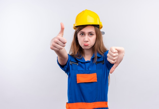 Młoda kobieta pracownik budowniczy w mundurze budowy i hełmie ochronnym pokazując kciuki w górę iw dół stojąc na białej ścianie