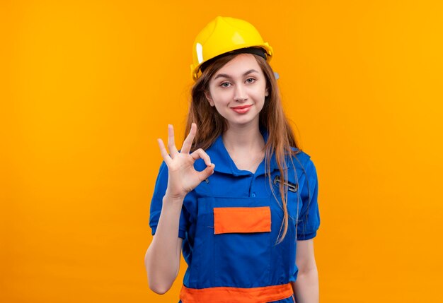 Młoda kobieta pracownik budowniczy w mundurze budowy i hełmie ochronnym uśmiechnięty pewny siebie robi ok znak stojący nad pomarańczową ścianą
